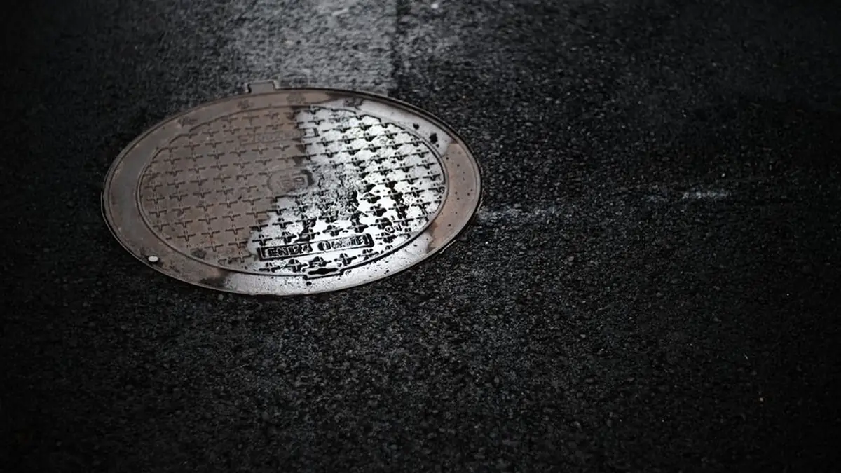 Wet cast metal access cover set into dark asphalt with water pooled across its surface