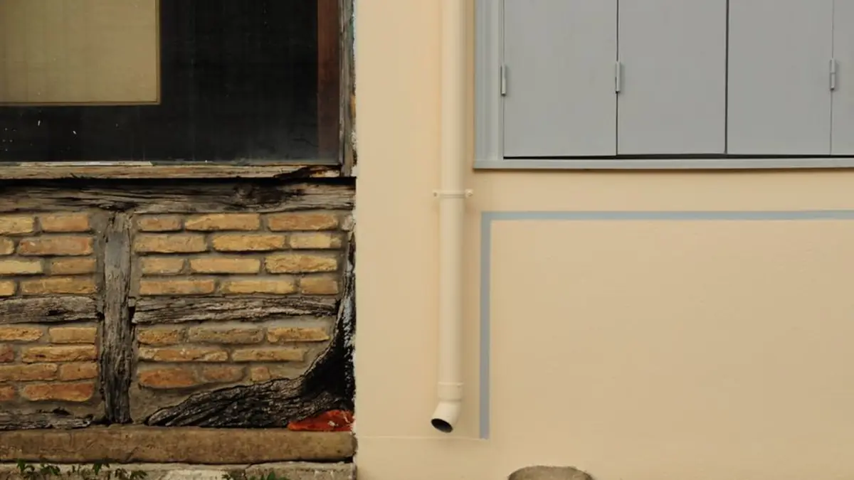 Weathered brick and timber wall beside a newly rendered one, with a white downpipe discharging at ground level
