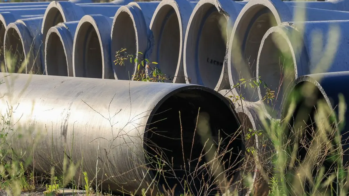 Rows of large concrete pipe sections stacked in a storage yard with grass growing around them