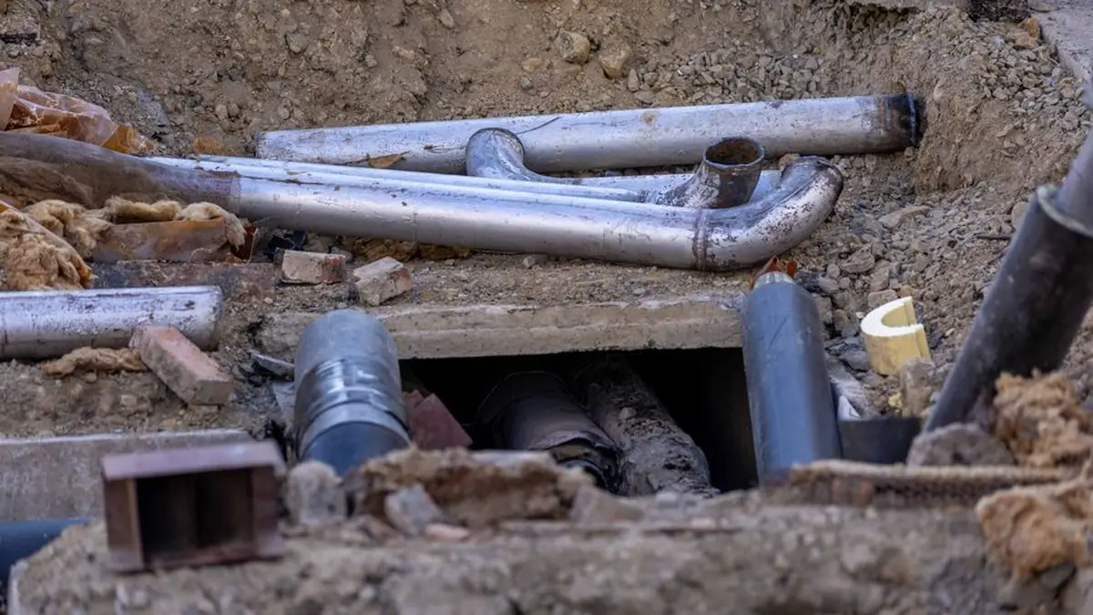 Old and new pipes exposed in an open excavation surrounded by broken brick, rubble and loose soil