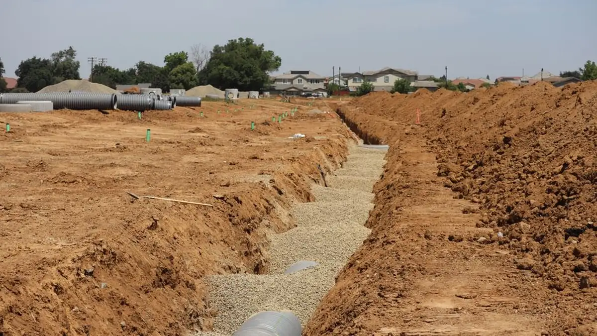 An open trench cut through orange soil with gravel bedding and pipe laid along the bottom