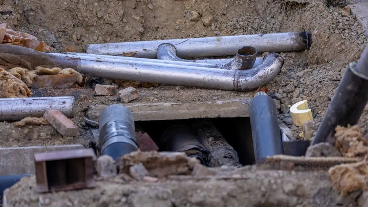 Old metal waste pipes and a bend exposed in an excavated trench beside a broken concrete slab