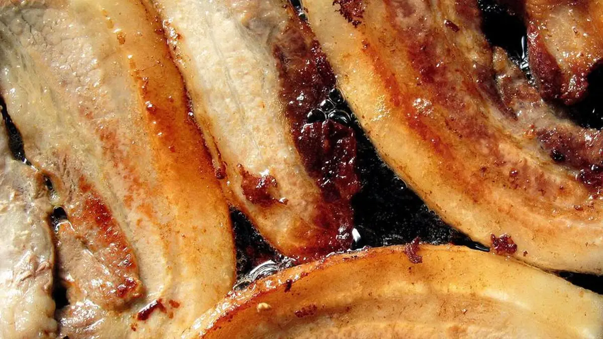 Close-up of rashers of bacon frying in their own melted fat in a dark pan