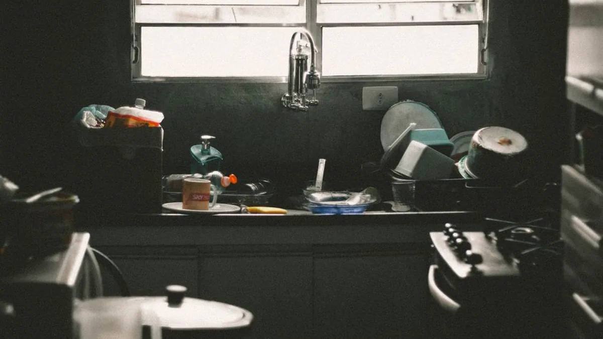 Dishes, pans and a dish rack stacked around a kitchen sink under a window in a dim kitchen