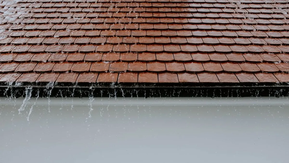Rain running down a brown tiled roof and dripping over the front edge of the gutter below it